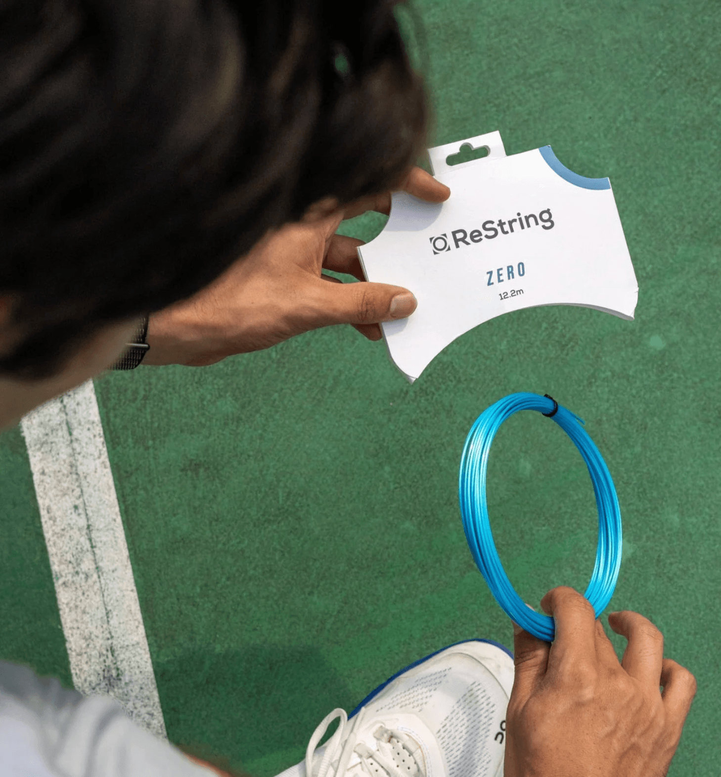 a person holding a set of restring zero tennis strings on a green tennis court