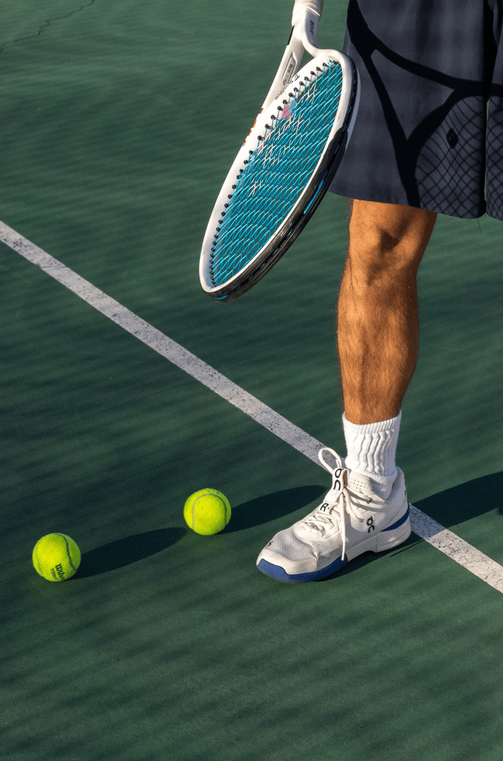 a person holding a racquet with blue strings on a tennis court with two tennis balls and ON tennis shoes