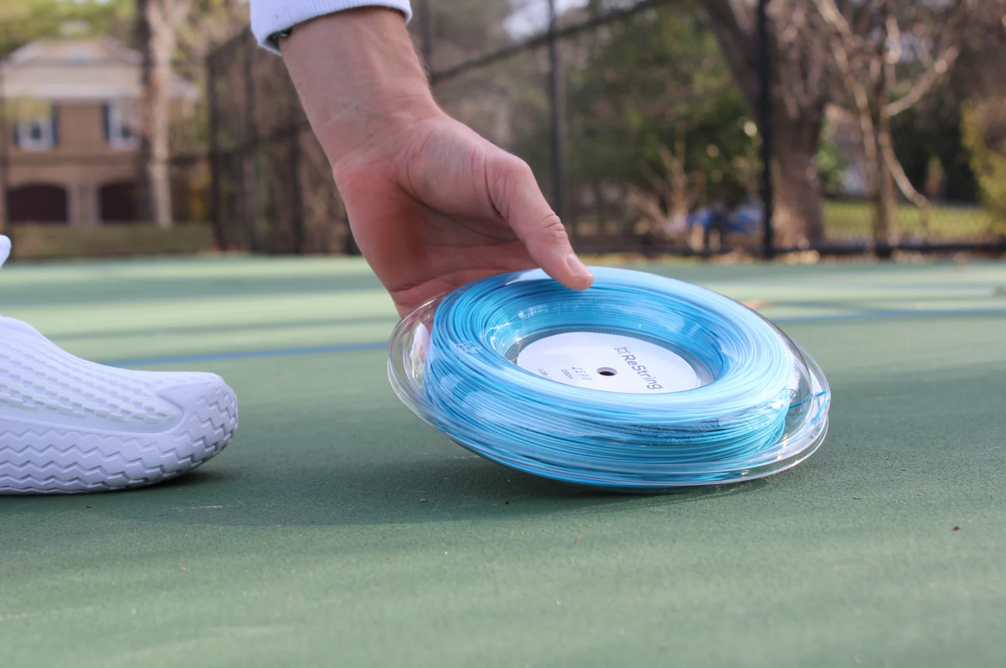 guy holding restring zero reel in blue colour on a green tennis court with trees in the background