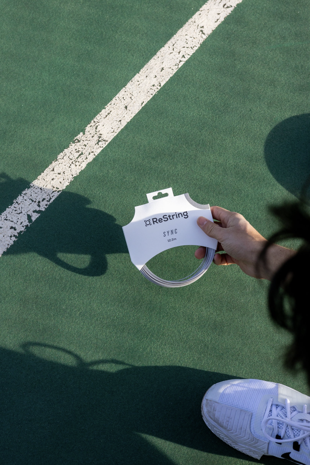 a man wearing cap holding a pack of restring sync on a tennis court